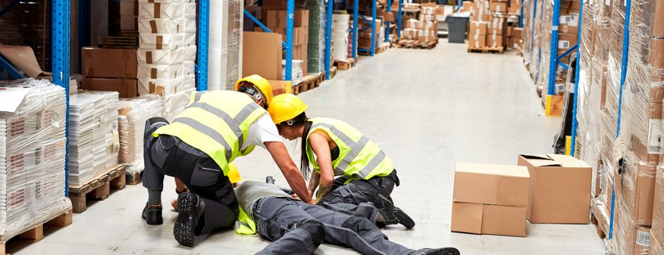 Accident in a warehouse with a member of staff laying on the floor with other employees crowding around the incident to help