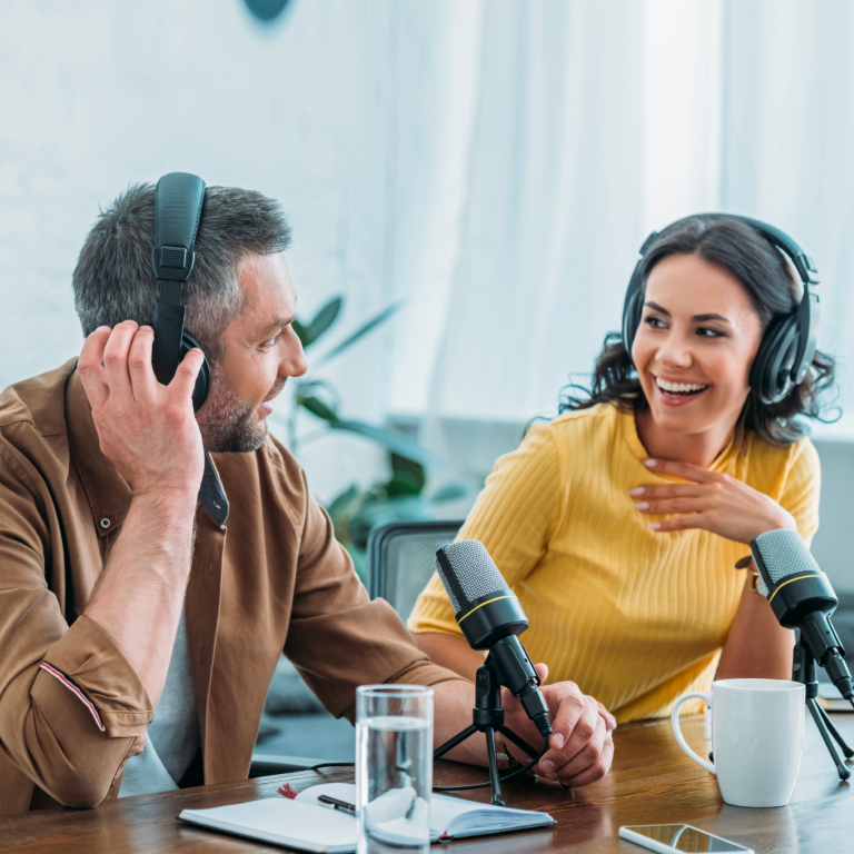 Podcast presenters laughing infront of microphones with plants in the background and a glass of water and a cup of coffee